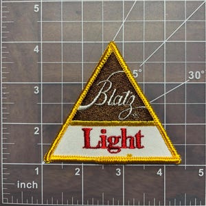May include: A triangular embroidered patch with a gold border. The patch features the words "Blatz Light" in white and red lettering on a white background. The top of the patch has a brown background with the word "Blatz" in white cursive lettering.