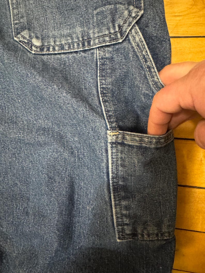 May include: Close-up of blue denim work pants with a pocket. The pocket is being held open by a hand. The denim has a worn appearance with visible stitching and a classic workwear aesthetic.