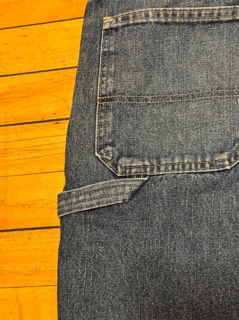 May include: Close-up of blue denim jeans, showcasing the back pocket and a utility pocket. The jeans have visible stitching and a classic design. The fabric appears worn, with a slight fade.