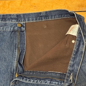 May include: Close-up of blue denim jeans with a brown fleece lining. The jeans have a button closure, a zipper, and a label visible on the inside. The denim is a medium wash, and the fleece lining is a warm brown color.