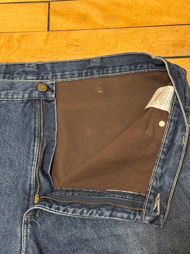 May include: Close-up of blue denim jeans with a brown fleece lining. The jeans have a button closure, a zipper, and a label visible on the inside. The denim is a medium wash, and the fleece lining is a warm brown color.