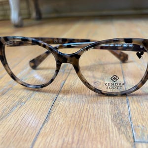May include: A pair of eyeglasses with a tortoiseshell pattern. The frames have a cat-eye shape and clear lenses. The brand name "Kendra Scott" is printed on the lens.