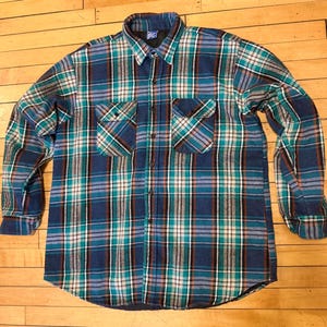 May include: A blue, green, and white plaid flannel shirt with two chest pockets. The long-sleeved shirt has a button-down front and a collar. The shirt is made of a thick, warm material.