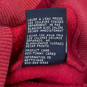 K&ouml;nnte beinhalten: Ein schwarzes Pflegeetikett mit wei&szlig;em Text in Englisch und Spanisch. Das Etikett enth&auml;lt Anweisungen zum Waschen und Trocknen des Kleidungsst&uuml;cks. Der Text lautet: "LAVER A L'EAU FROIDE LES COULEURS FONCEES SEPAREMENT. NE PAS BLANCHIR AVEC CHLORE SECHER IMMEDIATEMENT LAVAR CON AGUA FRIA LOS COLORES OSCUROS SEPARADO. NO BLANQUEADOR CLORADO SECAR PRONTO PRODUCT CARE: 800-TOMMY CARES INFORMATION DE NETTOYAGE: 800-866-6922"