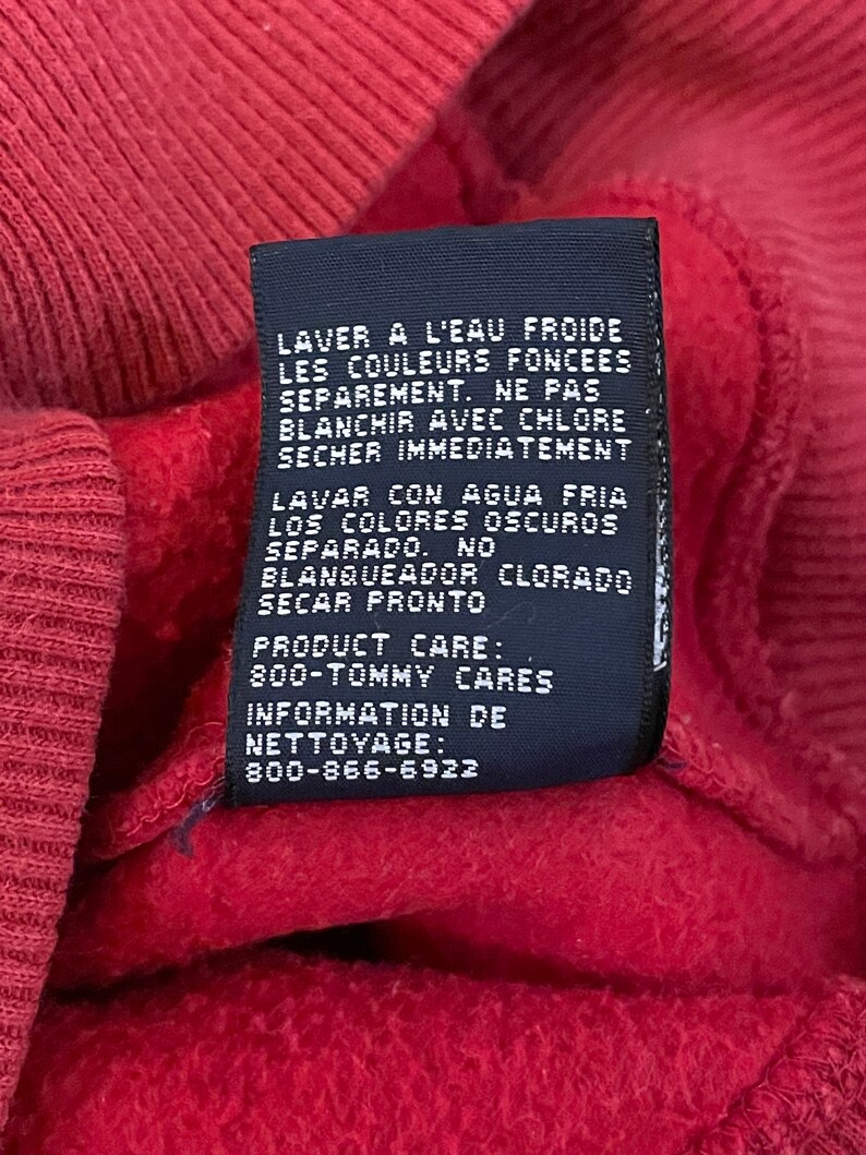 K&ouml;nnte beinhalten: Ein schwarzes Pflegeetikett mit wei&szlig;em Text in Englisch und Spanisch. Das Etikett enth&auml;lt Anweisungen zum Waschen und Trocknen des Kleidungsst&uuml;cks. Der Text lautet: "LAVER A L'EAU FROIDE LES COULEURS FONCEES SEPAREMENT. NE PAS BLANCHIR AVEC CHLORE SECHER IMMEDIATEMENT LAVAR CON AGUA FRIA LOS COLORES OSCUROS SEPARADO. NO BLANQUEADOR CLORADO SECAR PRONTO PRODUCT CARE: 800-TOMMY CARES INFORMATION DE NETTOYAGE: 800-866-6922"