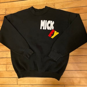 May include: Black sweatshirt with a white "MICK EY" graphic on the front. The letters are in a bold, blocky font. The "EY" is in red and yellow.
