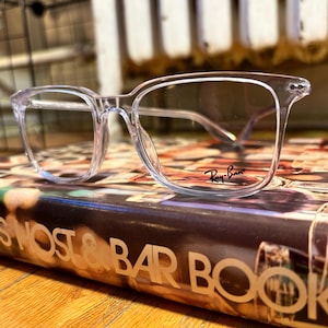 May include: Clear Ray-Ban eyeglasses with a square frame design. The glasses are resting on a book with the text "BOY'S MOST & BAR BOOK". The Ray-Ban logo is visible on the lens.