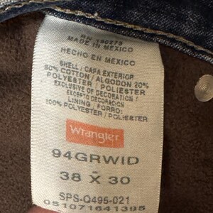 May include: Close-up of a Wrangler jeans tag. The tag is white with the brand name in orange. It lists the size as 38 x 30 inches, and the material composition as cotton and polyester. Made in Mexico.