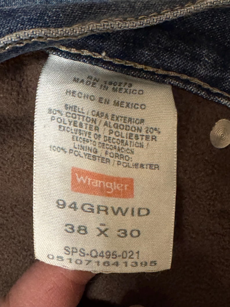 May include: Close-up of a Wrangler jeans tag. The tag is white with the brand name in orange. It lists the size as 38 x 30 inches, and the material composition as cotton and polyester. Made in Mexico.
