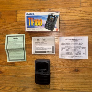 May include: A black Casio TV-100 pocket color television with a 1.6 inch screen, accompanied by its original retail box, an instruction manual, a warranty card, and a car or AC adaptor order form, all featuring the Casio brand name and logo.