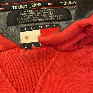 K&ouml;nnte beinhalten: Roter Kapuzenpullover mit einem schwarzen Tommy Jeans-Etikett. Das Etikett tr&auml;gt die Aufschrift "TOMMY JEANS" und "MADE IN PAKISTAN" mit dem Tommy-Flaggenlogo. Das Etikett enth&auml;lt auch die Stoffzusammensetzung: "80% COTTON/COTON/ALGODON 20% POLYESTER/POLIESTER".