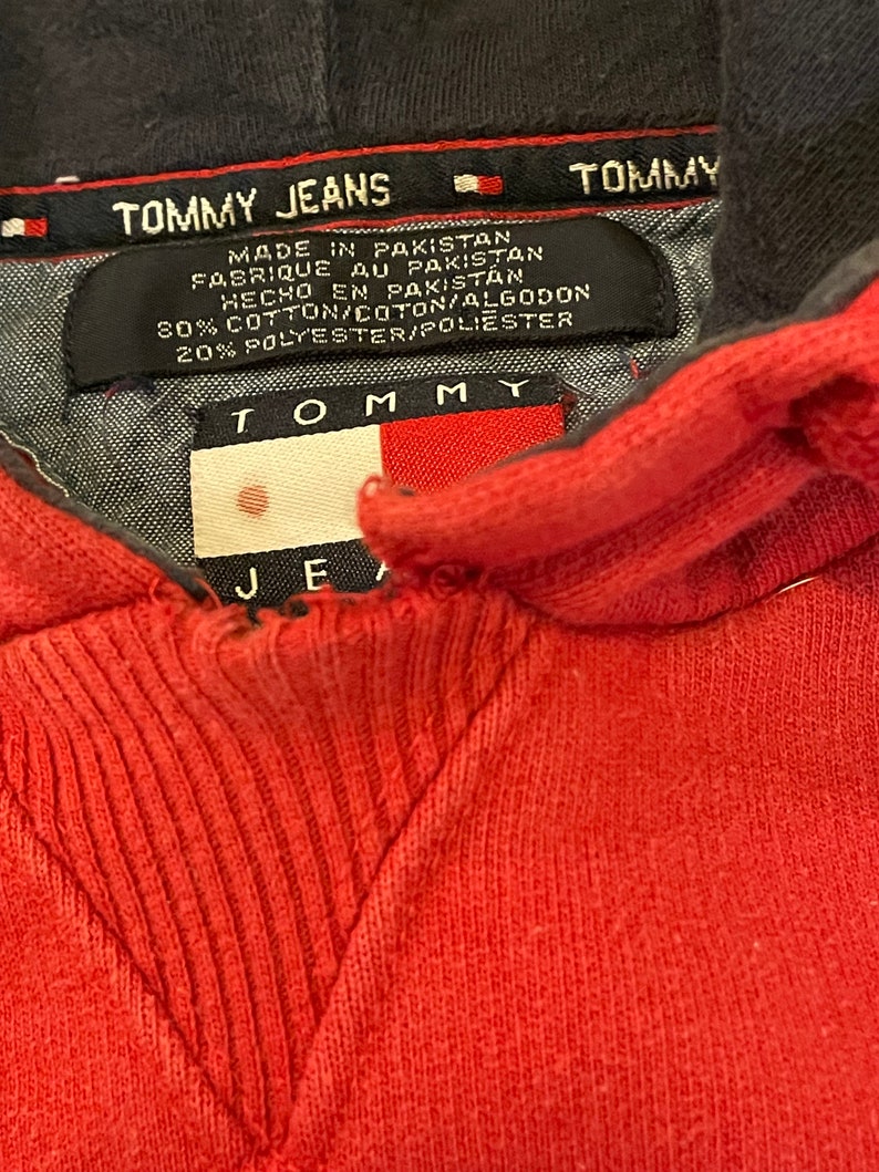 K&ouml;nnte beinhalten: Roter Kapuzenpullover mit einem schwarzen Tommy Jeans-Etikett. Das Etikett tr&auml;gt die Aufschrift "TOMMY JEANS" und "MADE IN PAKISTAN" mit dem Tommy-Flaggenlogo. Das Etikett enth&auml;lt auch die Stoffzusammensetzung: "80% COTTON/COTON/ALGODON 20% POLYESTER/POLIESTER".