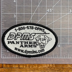 May include: White oval patch with black embroidered border. The patch features the text "DPMS", "PANTHER ARMS", and "www.dpmsinc.com". A white panther head is also embroidered on the patch.
