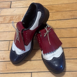 May include: A pair of tri-colored oxford shoes featuring navy blue leather toe caps and heels, white textured leather side panels, and deep red leather kiltie fringe overlays. The shoes include beige laces and decorative brogue detailing.