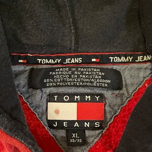 K&ouml;nnte beinhalten: Roter Kapuzenpullover mit einem schwarzen Tommy Jeans Logo und einem wei&szlig;-blauen Tommy Jeans Flaggenlogo. Das Etikett lautet "Hergestellt in Pakistan" und "80% Baumwolle/20% Polyester". Das Gr&ouml;&szlig;enetikett lautet "XL XG/X6".