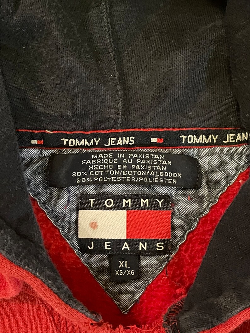 K&ouml;nnte beinhalten: Roter Kapuzenpullover mit einem schwarzen Tommy Jeans Logo und einem wei&szlig;-blauen Tommy Jeans Flaggenlogo. Das Etikett lautet "Hergestellt in Pakistan" und "80% Baumwolle/20% Polyester". Das Gr&ouml;&szlig;enetikett lautet "XL XG/X6".