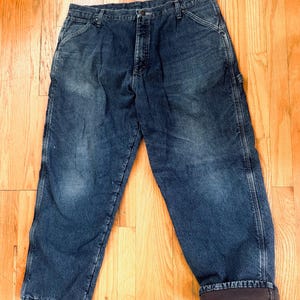 May include: Dark blue denim work pants with a faded appearance. The pants have a classic five-pocket design, a button and zipper closure, and a relaxed fit. The cuffs are rolled up, revealing a brown lining.