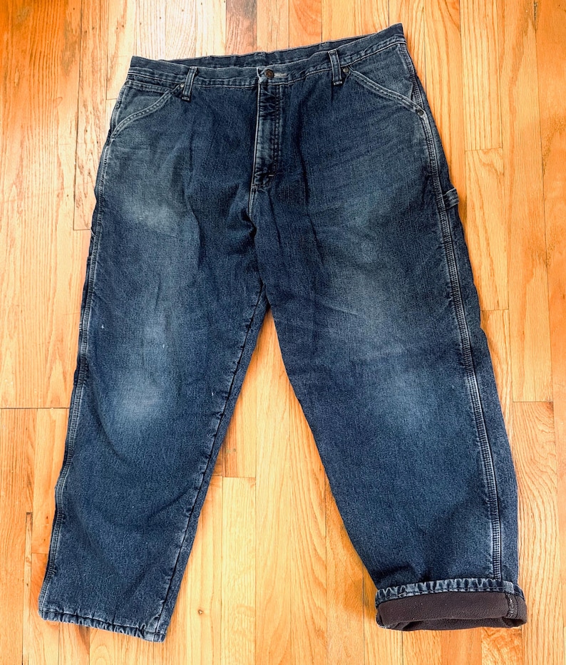 May include: Dark blue denim work pants with a faded appearance. The pants have a classic five-pocket design, a button and zipper closure, and a relaxed fit. The cuffs are rolled up, revealing a brown lining.