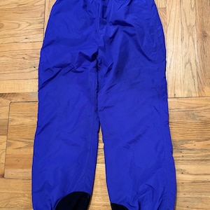 May include: Bright blue snow pants with an elastic waistband and black patches at the ankles. The pants are made of a lightweight, water-resistant material, perfect for winter activities. The pants are laid flat on a wooden surface.