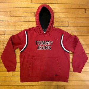 K&ouml;nnte beinhalten: Roter Kapuzenpullover mit wei&szlig;en und dunkelblauen Streifen an den &Auml;rmeln. Der Pullover tr&auml;gt den Schriftzug "TOMMY JEANS" in Schwarz, umrandet von Wei&szlig;.