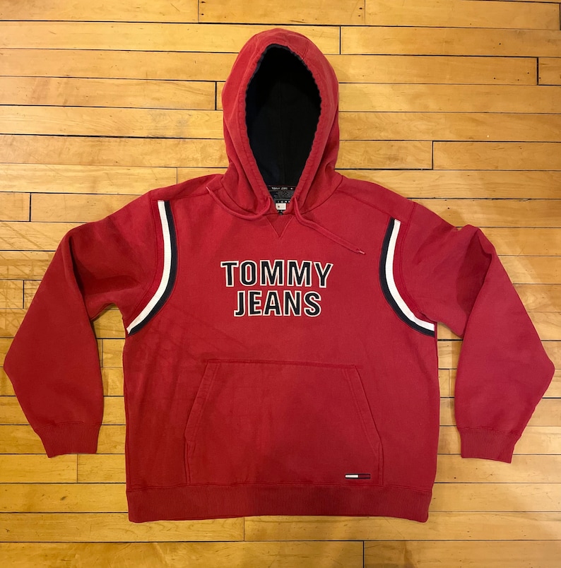 K&ouml;nnte beinhalten: Roter Kapuzenpullover mit wei&szlig;en und dunkelblauen Streifen an den &Auml;rmeln. Der Pullover tr&auml;gt den Schriftzug "TOMMY JEANS" in Schwarz, umrandet von Wei&szlig;.
