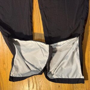 May include: Black athletic pants with zippers at the ankles, revealing a light gray lining. The pants are made of a smooth, possibly nylon or polyester material, and are laid out on a wooden surface.