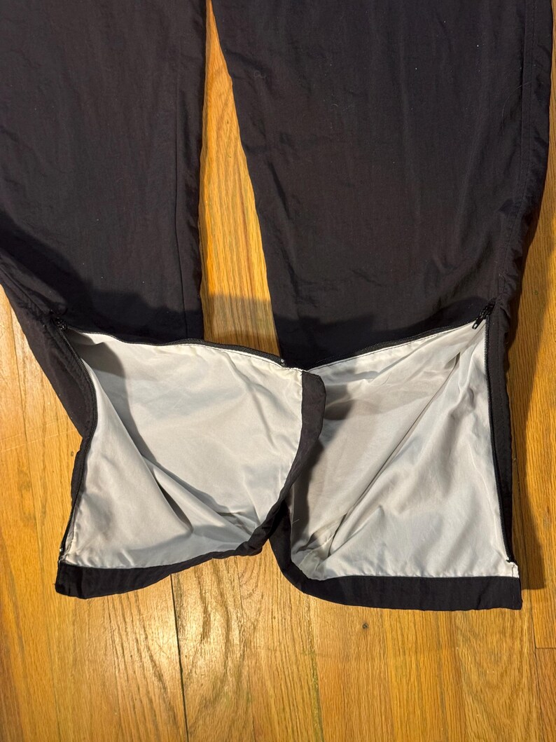 May include: Black athletic pants with zippers at the ankles, revealing a light gray lining. The pants are made of a smooth, possibly nylon or polyester material, and are laid out on a wooden surface.