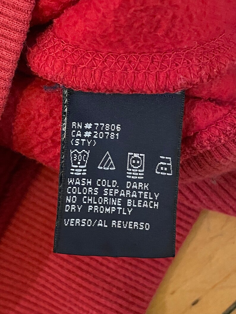 K&ouml;nnte beinhalten: Ein dunkelblaues Pflegeetikett mit wei&szlig;em Text und Symbolen. Der Text lautet "RN #77806 CA#20781 (STY) WASH COLD. DARK COLORS SEPARATELY NO CHLORINE BLEACH DRY PROMPTLY VERSO/AL REVERSO".