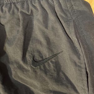 May include: Close-up of black athletic pants with the Nike swoosh logo. The pants have an elastic waistband and a pocket detail. The fabric appears to be a lightweight, slightly shiny material.