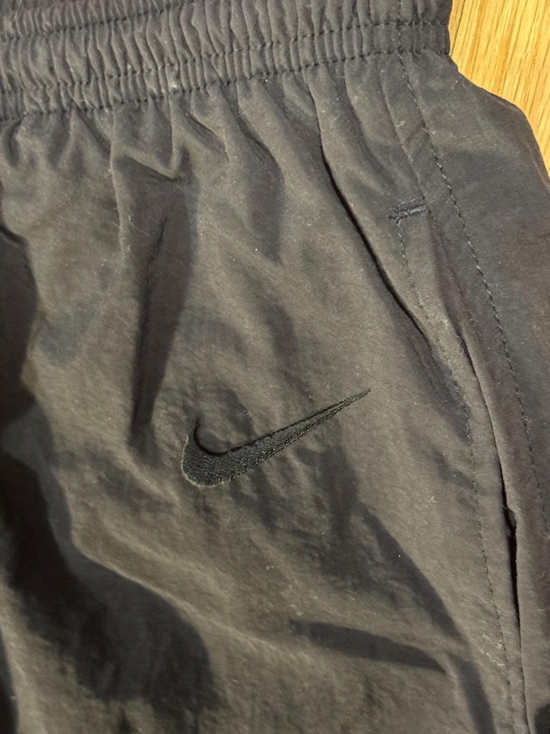 May include: Close-up of black athletic pants with the Nike swoosh logo. The pants have an elastic waistband and a pocket detail. The fabric appears to be a lightweight, slightly shiny material.