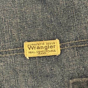 May include: Close-up of blue denim fabric with a rectangular tan label. The label reads "AUTHENTIC ISSUE WRANGLER REAL COMFORTABLE JEANS" in a bold, serif font. The denim has a textured appearance.