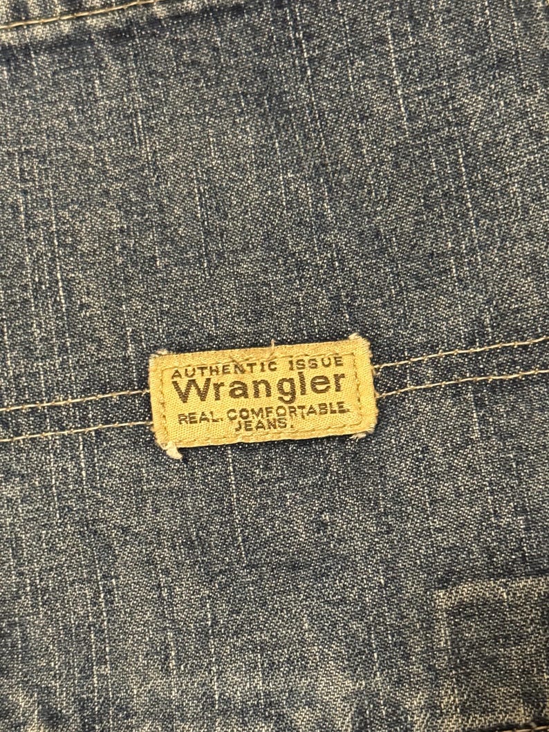 May include: Close-up of blue denim fabric with a rectangular tan label. The label reads "AUTHENTIC ISSUE WRANGLER REAL COMFORTABLE JEANS" in a bold, serif font. The denim has a textured appearance.