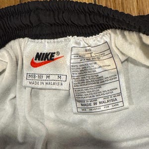May include: Close-up of a white and black Nike athletic shorts tag. The tag includes the Nike logo, size information, and material composition details. The shorts are made in Malaysia.