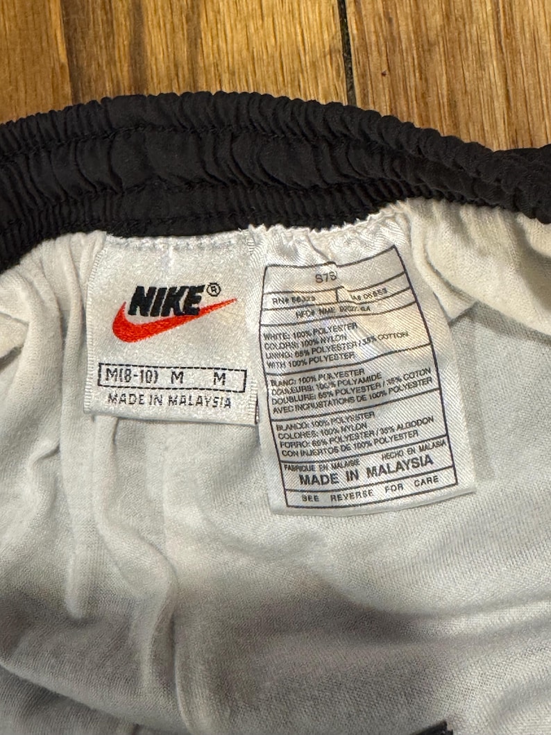 May include: Close-up of a white and black Nike athletic shorts tag. The tag includes the Nike logo, size information, and material composition details. The shorts are made in Malaysia.