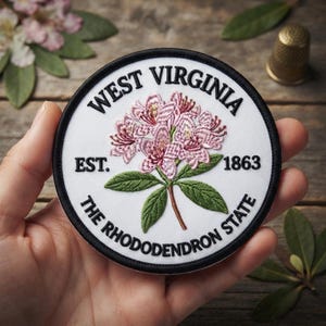 May include: A circular embroidered patch with a white background and a black border. The center features a pink rhododendron flower with green leaves. Text reads: WEST VIRGINIA, EST. 1863, and THE RHODODENDRON STATE in black capital letters.