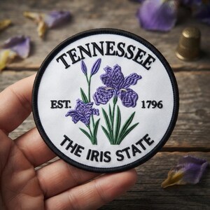 May include: A circular embroidered patch with a white background and a black border. It features purple iris flowers with green stems and leaves. The text reads TENNESSEE, EST. 1796, and THE IRIS STATE in black capital letters around the floral design.