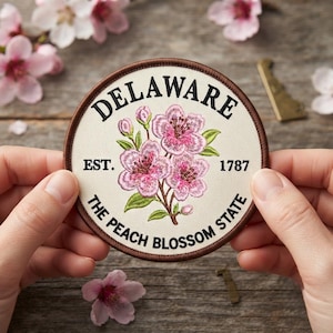 May include: A circular embroidered patch with a cream-colored background and a dark brown border. The text reads DELAWARE, EST. 1787, and THE PEACH BLOSSOM STATE. In the center, there is an embroidered illustration of pink peach blossoms and green leaves.