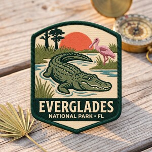 May include: An embroidered patch featuring a green alligator in blue water, a pink roseate spoonbill, and a red sun over a landscape. The text reads EVERGLADES NATIONAL PARK FL in cream letters on a dark green background with a dark green border.