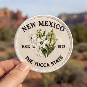 May include: A circular embroidered patch with a cream-colored background featuring a central illustration of white yucca flowers and green leaves. The text reads NEW MEXICO, EST. 1912, and THE YUCCA STATE in black, stitched lettering.