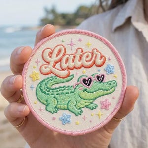 May include: A circular embroidered patch with a cream-colored background and a pink stitched border. The patch features a green alligator wearing pink heart-shaped sunglasses, surrounded by small colorful stars and the word Later in orange script.