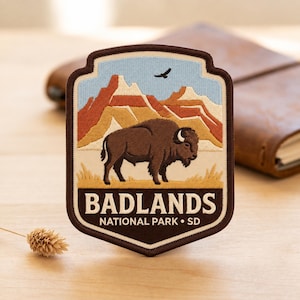 May include: A shield-shaped embroidered patch featuring a brown bison standing in a grassy field with red and tan rock formations in the background under a light blue sky with a bird. The text reads BADLANDS NATIONAL PARK • SD in white.