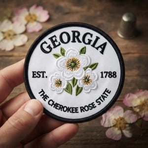 May include: A circular embroidered patch with a white background and black border. It features three white Cherokee Rose flowers with green leaves in the center. The text reads GEORGIA, EST. 1788, and THE CHEROKEE ROSE STATE in black lettering.