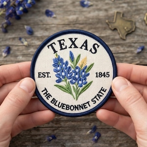 May include: A circular embroidered patch with a cream-colored fabric background and a dark blue border. The design features bluebonnet flowers in blue and green with a yellow accent, the text TEXAS, EST. 1845, and THE BLUEBONNET STATE.