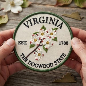 May include: A circular embroidered patch featuring a cream-colored background with a green border. The text reads VIRGINIA, EST. 1788, and THE DOGWOOD STATE. A central illustration depicts a branch with white dogwood flowers and green leaves.