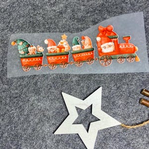 Könnte beinhalten: Eine weihnachtliche Illustration eines Zuges mit Weihnachtsmann und Wichteln in roter und grüner Kleidung. Die Waggons sind rot mit orangefarbenen Rädern. Ein weißer Stern und Holzelemente sind ebenfalls vorhanden.