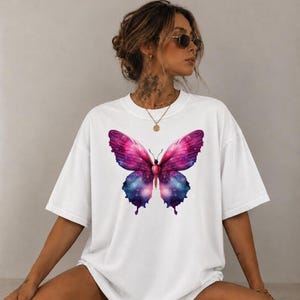 Könnte beinhalten: Ein weißes T-Shirt mit kurzen Ärmeln und einer großen, mittigen Grafik eines Schmetterlings, dessen Flügel ein lebhaftes Galaxie-Muster in dunklen Rosa-, Violett- und Blautönen zeigen, akzentuiert durch kleine weiße Punkte, die an ferne Sterne erinnern.