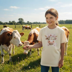 Könnte beinhalten: Beiges T-Shirt mit einer Cartoon-Kuh mit Blumenkranz. Die Kuh hat ein weißes Gesicht, braune Hörner und rosa Blumen. Das T-Shirt wird von einer Person auf einer grünen Wiese mit grasenden Kühen im Hintergrund getragen.