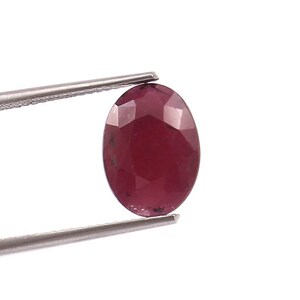 May include: An oval-shaped, faceted ruby gemstone held by silver tweezers. The ruby has a deep red color with visible internal inclusions. The gem is set against a plain white background.