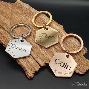 Può includere: Tre portachiavi esagonali in oro, argento e oro rosa con i nomi incisi "Pepe", "Simon" e "Odin". Ogni portachiavi presenta un elemento di design unico: un motivo floreale, impronte di zampe e uccelli in volo.