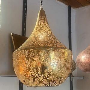Puede incluir: Lámpara colgante de latón con forma de lágrima. La lámpara tiene un intrincado patrón de pequeños agujeros que permiten que la luz brille. Esta lámpara decorativa está suspendida de una cadena.
